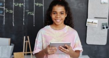 garota negra de cabelos longos sorri enqquanto segura um tablet na frente de um quadro negro (palavras de destaque: educação básica)