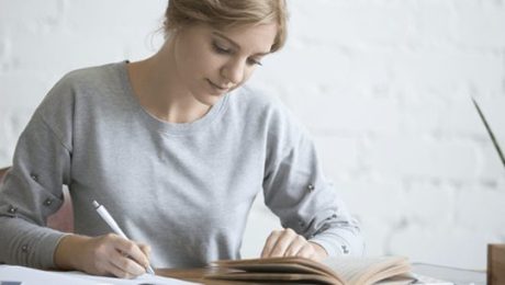 mulher de pele clara e cabelos presos escreve numa folha enquanto lê. Esse conteúdo fala sobre educação.
