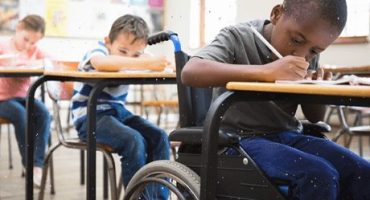 menino em cadeira de rodas escreve na sua carteira e colegas na fila detrás fazem o mesmo - esse conteúdo trata de educação formal