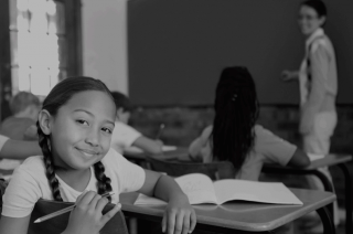 foto preta e branca de crianças em sala de aula e uma dela olha para a câmera.