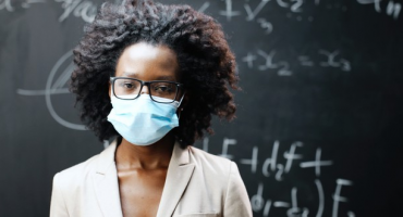 professora negra de óculos usa máscara de proteção à frente do quadro negro