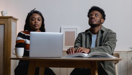 Sentados à mesa, homem negro cego lê braille ao lado de mulher negra que usa computador