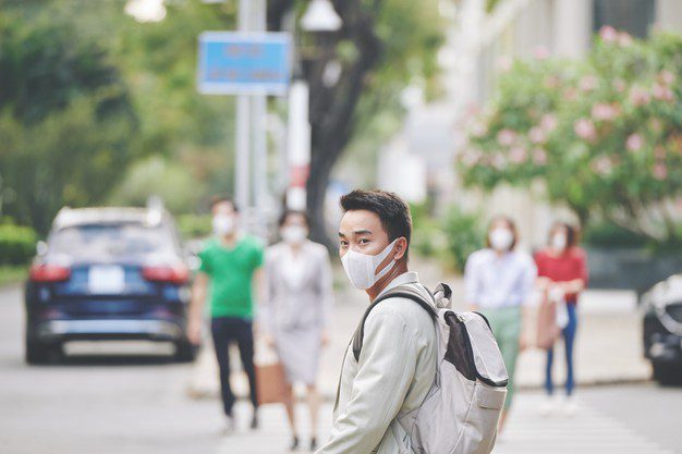 Homem oriental e de máscara no rosto usando mochila e atravessando a rua para ir à escola