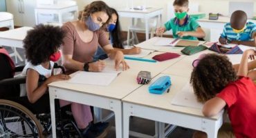 Foto de professora sentada a uma grande mesa com alunos ao redor, todos usam máscara cirúrgica. Uma aluna está em cadeira de rodas.