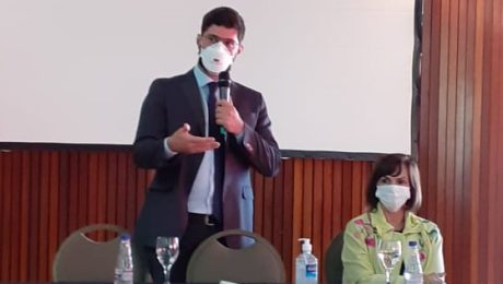 Foto de uma bancada, com Lucas Hoogerbrugge, homem, branco, usando máscara cirúrgica, de pé falando ao microfone. Uma mulher está sentada ao seu lado.