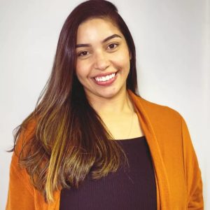 Doane, mulher branca de cabelos castanhos longos. Ela sorri e veste casaco laranja sobre blusa preta
