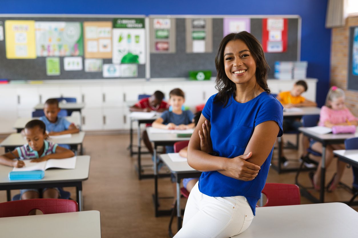 Professora negra sorri de braços cruzados com sala e aula com alunos ao fundo
