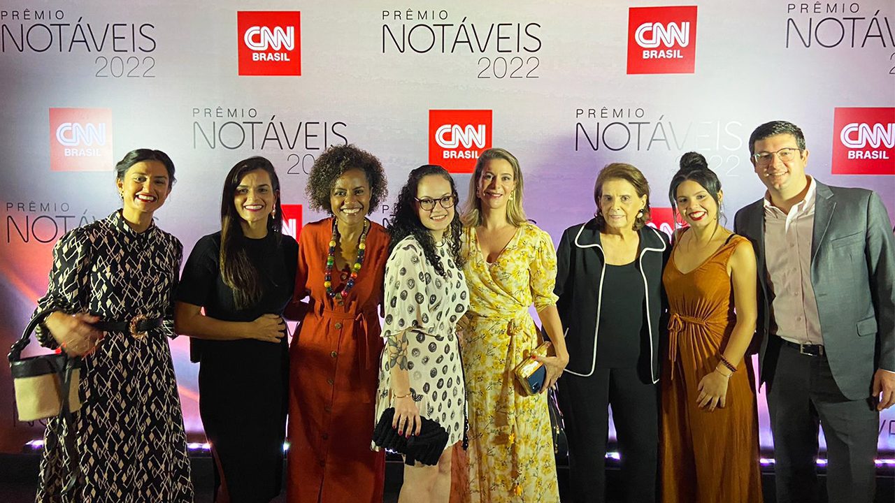 Sete mulheres diversas e um homem sorriem para a foto, em frente ao painel Prêmio CNN Notáveis 2022.