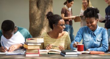 Três alunos negros sentados em uma mesa, estudando com livros ao lado.