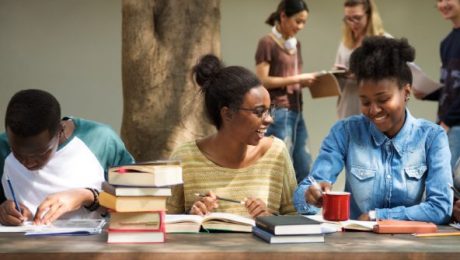 Três alunos negros sentados em uma mesa, estudando com livros ao lado.