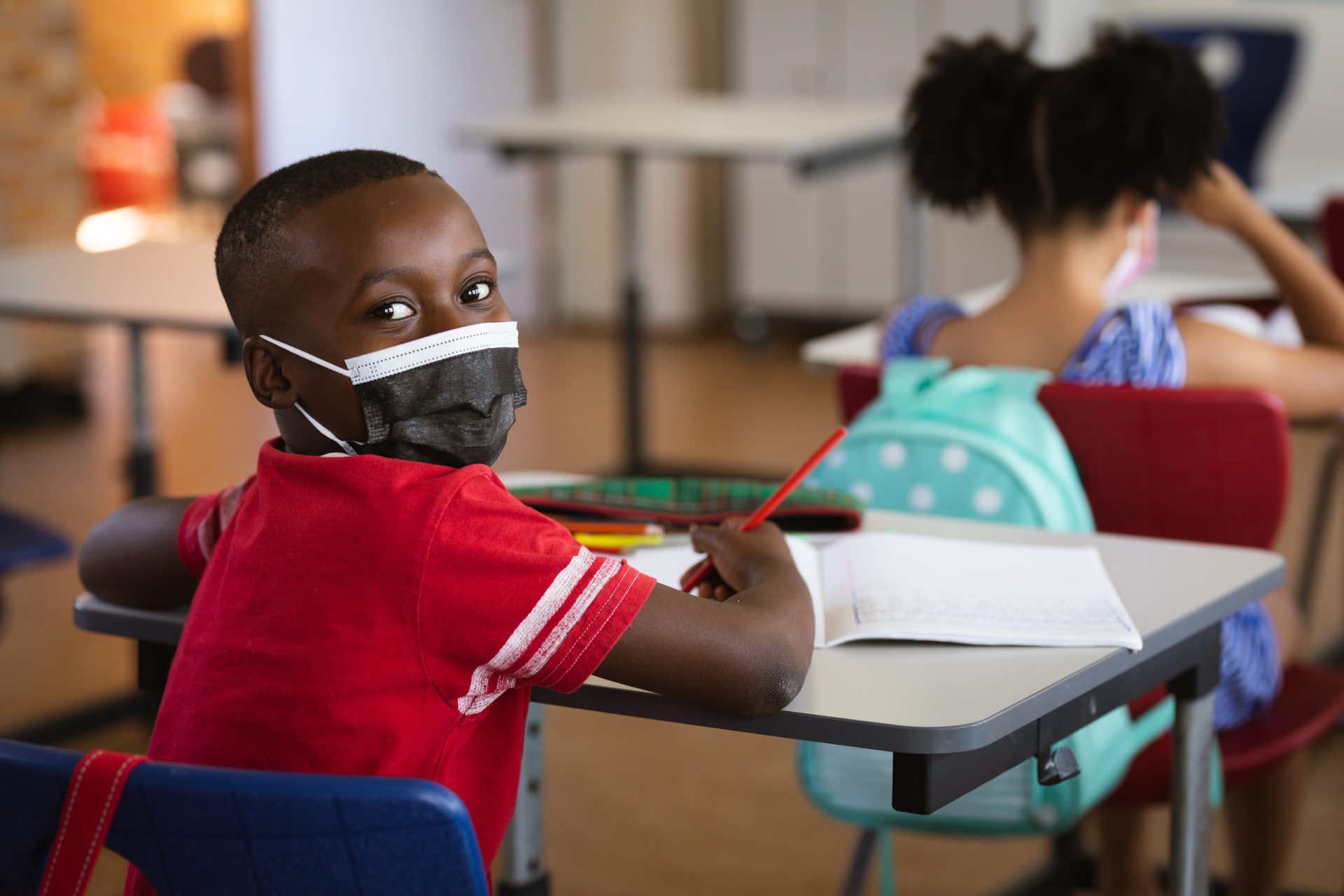 Imagem de uma criança negra em uma sala de aula, usando máscara e segurando um lápis