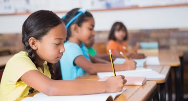 Meninas enfileiradas lado a lado em sala de aula, escrevendo com lápis na mão