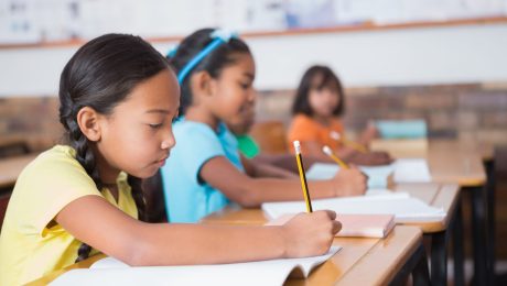 Meninas enfileiradas lado a lado em sala de aula, escrevendo com lápis na mão