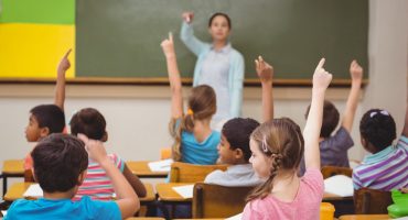Professora a frente da sala de aula e alunos sentados, de costas e erguendo as mãos para participar da aula
