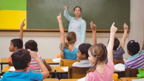 Professora a frente da sala de aula e alunos sentados, de costas e erguendo as mãos para participar da aula