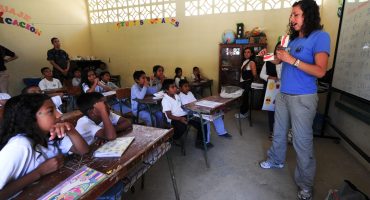 Foto de uma sala de aula com a professora explicando algo para os alunos que prestam atenção nela.
