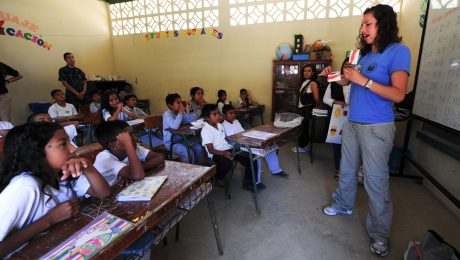 Foto de uma sala de aula com a professora explicando algo para os alunos que prestam atenção nela.