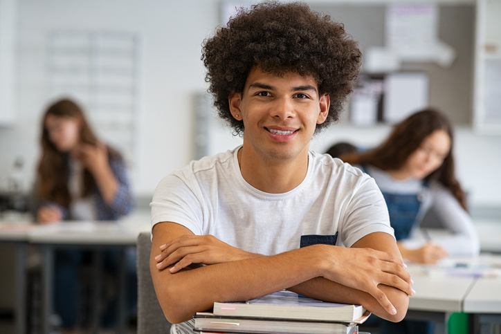 Garoto negro, de cabelos cacheados, sentado em sala de aula, com camiseta branca, sorrindo, de braços cruzados em cima de livros e olhando fixamente para frente