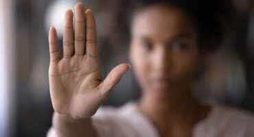Mulher negra em foco mostrando a palma da mão, em um sinal de parar. Ela está com camiseta preta e olhar combativo