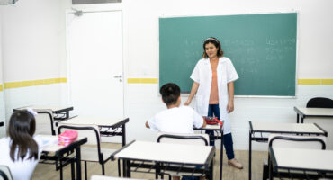 professora em sala de aula com 2 crianças sentadas de costas