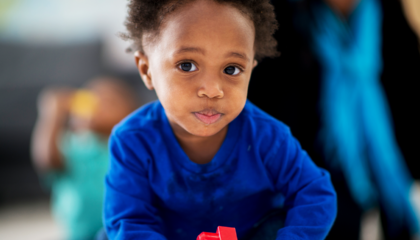 Menina de aproximadamente 3 anos e negro olha para câmera fazendo bico. Ele veste camisa de manga longa azul e está no ambiente escolar