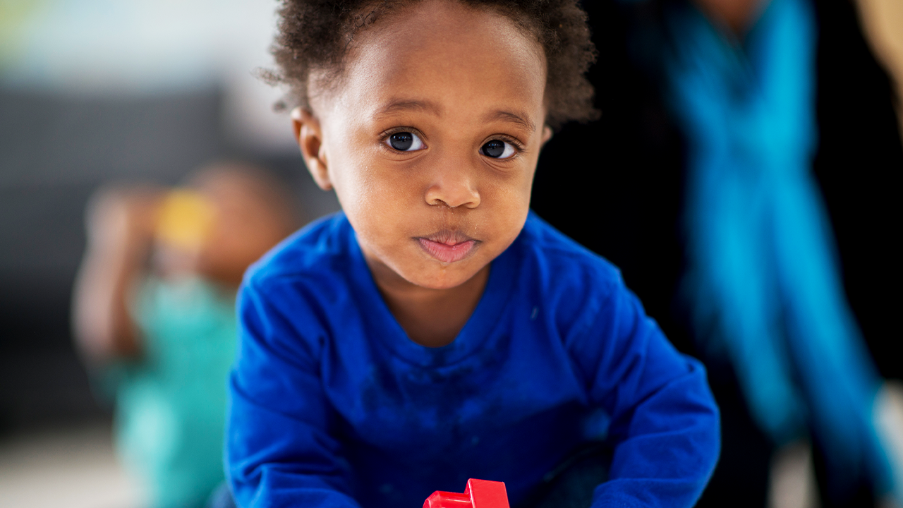 Menina de aproximadamente 3 anos e negro olha para câmera fazendo bico. Ele veste camisa de manga longa azul e está no ambiente escolar