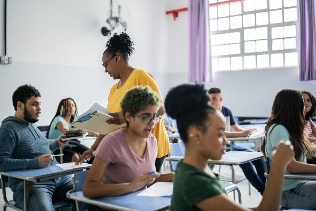 ambiente de sala de aula com estudantes adultos sentados em carteiras enquanto trabalham em materiais de estudo. Uma professora está de pé entre as carteiras, segurando alguns papéis e um livro, parecendo estar em meio a uma explicação ou discussão com a turma. Os alunos parecem atentos e engajados, com alguns deles escrevendo em cadernos. A sala é iluminada, com janelas grandes que permitem a entrada de luz natural, e as paredes são claras, o que contribui para uma atmosfera de aprendizado aberta e acolhedora.