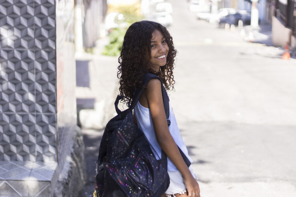 A imagem mostra uma jovem em um ambiente externo ensolarado. Ela está sorrindo e olhando para trás, por cima do ombro, com uma expressão relaxada e alegre. Seus cabelos são encaracolados e ela está vestindo um camiseta sem mangas, shorts e uma mochila, o que sugere um estilo casual e confortável, possivelmente indo para a escola. O fundo inclui uma rua que parece tranquila, com carros estacionados ao lado, e um edifício com uma parede de padrão geométrico, adicionando uma estética moderna à cena.