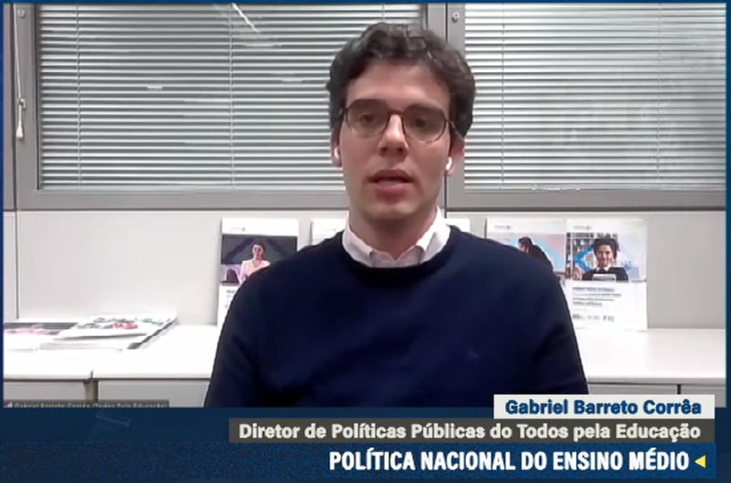 Na imagem, há um homem vestindo um suéter azul escuro sobre uma camisa de colarinho branco. Ele está usando óculos e parece estar falando ou apresentando algo. Atrás dele, há uma janela com persianas horizontais meio fechadas. Há também uma parede branca com cartazes que incluem textos e imagens de pessoas, possivelmente materiais informativos ou promocionais. Na parte inferior da imagem, há uma legenda em português que identifica o homem como "Gabriel Barreto Corrêa", e o título "Diretor de Políticas Públicas do Todos pela Educação". A legenda também menciona "POLÍTICA NACIONAL DO ENSINO MÉDIO", indicando que o assunto tratado pode estar relacionado a políticas educacionais no Brasil.