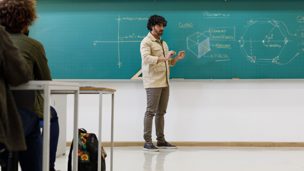 Professor em uma sala de aula explicando conteúdo que aparece na lousa.