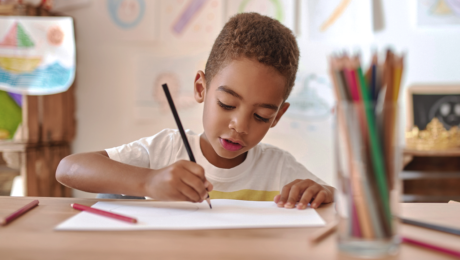 criança parda de cabelos castanhos claros e curtos escreve com lápis numa folha