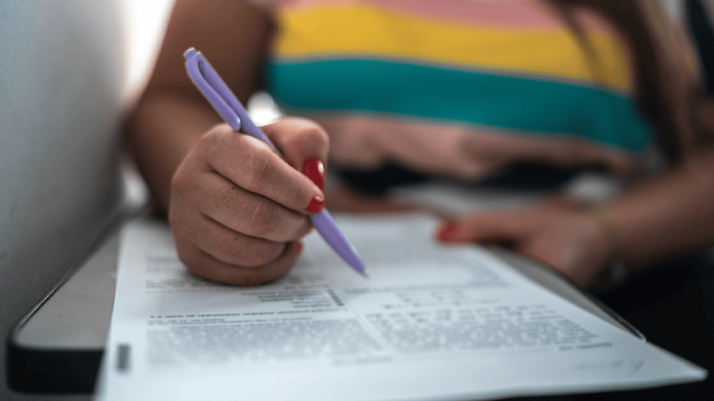 Com fundo desfocado, detalhe das mãos de uma pessoa segurando uma caneta roxa enquanto preenche uma folha de papel. Apenas a mão e parte do braço estão visíveis. As unhas estão pintadas de vermelho.