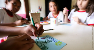 Recomendações para enfrentar os impactos da crise climática na Educação. Três crianças estão sentadas em volta de uma mesa branca, participando de uma atividade de desenho em um ambiente interno, provavelmente uma sala de aula. No primeiro plano, uma mão segura um lápis verde, desenhando flores, um sol e outros elementos coloridos em um papel azul-claro. Ao fundo, as crianças, que vestem camisetas brancas com detalhes em vermelho, estão concentradas, observando ou realizando suas próprias atividades. Há materiais escolares, como canetas e lápis de cor, espalhados sobre a mesa. A iluminação natural destaca o foco na criatividade e no aprendizado.