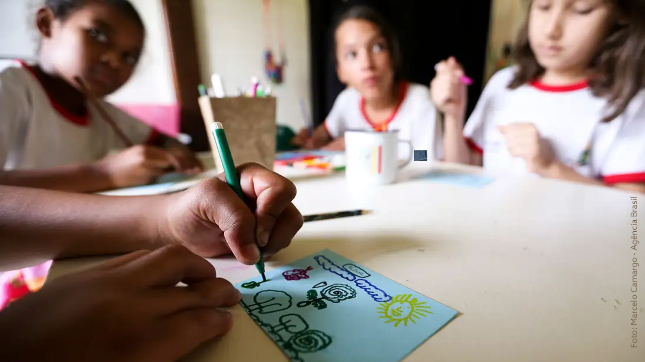 Recomendações para enfrentar os impactos da crise climática na Educação. Três crianças estão sentadas em volta de uma mesa branca, participando de uma atividade de desenho em um ambiente interno, provavelmente uma sala de aula. No primeiro plano, uma mão segura um lápis verde, desenhando flores, um sol e outros elementos coloridos em um papel azul-claro. Ao fundo, as crianças, que vestem camisetas brancas com detalhes em vermelho, estão concentradas, observando ou realizando suas próprias atividades. Há materiais escolares, como canetas e lápis de cor, espalhados sobre a mesa. A iluminação natural destaca o foco na criatividade e no aprendizado.