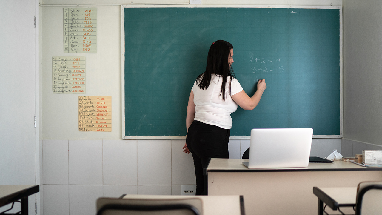 Uma mulher de cabelos longos e escuros está em pé, de costas, escrevendo com giz em um quadro verde de sala de aula. Ela veste uma camiseta branca e calça preta. No quadro, há operações matemáticas escritas: 