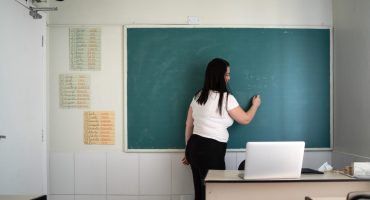 Uma mulher de cabelos longos e escuros está em pé, de costas, escrevendo com giz em um quadro verde de sala de aula. Ela veste uma camiseta branca e calça preta. No quadro, há operações matemáticas escritas: 