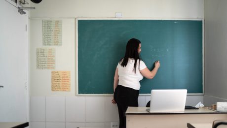 Uma mulher de cabelos longos e escuros está em pé, de costas, escrevendo com giz em um quadro verde de sala de aula. Ela veste uma camiseta branca e calça preta. No quadro, há operações matemáticas escritas: 