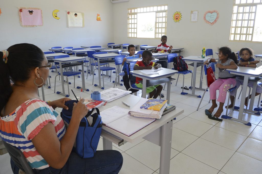 A imagem retrata uma sala de aula em uma escola pública, onde uma professora e um pequeno grupo de crianças estão presentes. A professora, que tem cabelo preso e usa óculos, está sentada em sua mesa, organizando materiais e escrevendo em um caderno. Sobre a mesa, há livros, cadernos, lápis e outros itens escolares. As crianças estão espalhadas por algumas carteiras, que são azuis e organizadas em filas. Algumas delas estão escrevendo ou desenhando em seus cadernos, enquanto outras parecem distraídas ou conversando. Duas meninas, sentadas juntas no canto direito da imagem, observam o ambiente, e uma delas está descalça. A sala de aula é iluminada pela luz natural que entra pelas janelas e tem paredes decoradas com cartazes coloridos e desenhos, incluindo um sol sorridente e recortes em forma de lua e estrela. No fundo, há várias carteiras vazias, sugerindo que a turma pode estar incompleta ou que nem todos os alunos compareceram naquele dia. A cena transmite um ambiente de aprendizado, mas também destaca desafios como a frequência escolar e a infraestrutura.