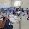 A imagem retrata uma sala de aula em uma escola pública, onde uma professora e um pequeno grupo de crianças estão presentes. A professora, que tem cabelo preso e usa óculos, está sentada em sua mesa, organizando materiais e escrevendo em um caderno. Sobre a mesa, há livros, cadernos, lápis e outros itens escolares. As crianças estão espalhadas por algumas carteiras, que são azuis e organizadas em filas. Algumas delas estão escrevendo ou desenhando em seus cadernos, enquanto outras parecem distraídas ou conversando. Duas meninas, sentadas juntas no canto direito da imagem, observam o ambiente, e uma delas está descalça. A sala de aula é iluminada pela luz natural que entra pelas janelas e tem paredes decoradas com cartazes coloridos e desenhos, incluindo um sol sorridente e recortes em forma de lua e estrela. No fundo, há várias carteiras vazias, sugerindo que a turma pode estar incompleta ou que nem todos os alunos compareceram naquele dia. A cena transmite um ambiente de aprendizado, mas também destaca desafios como a frequência escolar e a infraestrutura.