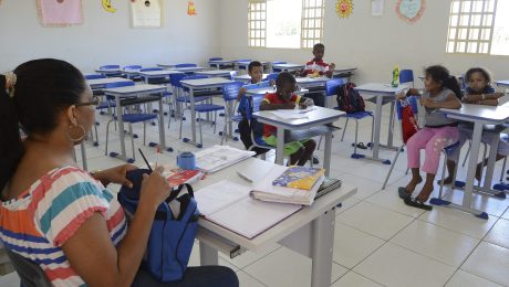 A imagem retrata uma sala de aula em uma escola pública, onde uma professora e um pequeno grupo de crianças estão presentes. A professora, que tem cabelo preso e usa óculos, está sentada em sua mesa, organizando materiais e escrevendo em um caderno. Sobre a mesa, há livros, cadernos, lápis e outros itens escolares. As crianças estão espalhadas por algumas carteiras, que são azuis e organizadas em filas. Algumas delas estão escrevendo ou desenhando em seus cadernos, enquanto outras parecem distraídas ou conversando. Duas meninas, sentadas juntas no canto direito da imagem, observam o ambiente, e uma delas está descalça. A sala de aula é iluminada pela luz natural que entra pelas janelas e tem paredes decoradas com cartazes coloridos e desenhos, incluindo um sol sorridente e recortes em forma de lua e estrela. No fundo, há várias carteiras vazias, sugerindo que a turma pode estar incompleta ou que nem todos os alunos compareceram naquele dia. A cena transmite um ambiente de aprendizado, mas também destaca desafios como a frequência escolar e a infraestrutura.