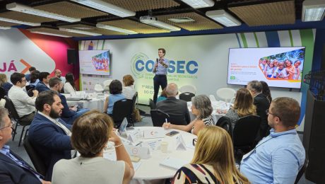 A imagem mostra um evento organizado pelo Conselho Nacional de Secretários das Capitais (Consec) em um ambiente moderno e bem iluminado. Vários participantes, incluindo gestores e especialistas em educação, estão reunidos ao redor de mesas dispostas em formato de conferência. No centro da imagem, um palestrante está de pé, segurando um microfone e apresentando informações projetadas em duas telas grandes ao fundo. Nas telas, há um slide com o título 