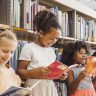 A imagem mostra quatro crianças em uma biblioteca, cercadas por prateleiras repletas de livros. Elas estão em pé, segurando livros abertos e sorrindo enquanto leem. A primeira criança, à esquerda, é uma menina loira de vestido laranja claro, olhando para seu livro com um sorriso suave. Ao lado dela, uma menina com cabelo cacheado preso em um coque alto veste uma blusa branca e uma saia clara, segurando um livro de capa vermelha e rindo. A terceira criança, uma menina de cabelos volumosos e cacheados, usa um vestido cinza com detalhe brilhante no peito e também segura um livro vermelho, sorrindo animada. Por fim, no canto direito, um menino de cabelos curtos e camisa clara está segurando um livro e rindo. O ambiente é iluminado, e o cenário transmite alegria e interesse pela leitura.