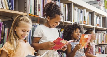 A imagem mostra quatro crianças em uma biblioteca, cercadas por prateleiras repletas de livros. Elas estão em pé, segurando livros abertos e sorrindo enquanto leem. A primeira criança, à esquerda, é uma menina loira de vestido laranja claro, olhando para seu livro com um sorriso suave. Ao lado dela, uma menina com cabelo cacheado preso em um coque alto veste uma blusa branca e uma saia clara, segurando um livro de capa vermelha e rindo. A terceira criança, uma menina de cabelos volumosos e cacheados, usa um vestido cinza com detalhe brilhante no peito e também segura um livro vermelho, sorrindo animada. Por fim, no canto direito, um menino de cabelos curtos e camisa clara está segurando um livro e rindo. O ambiente é iluminado, e o cenário transmite alegria e interesse pela leitura.