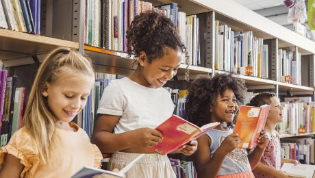 A imagem mostra quatro crianças em uma biblioteca, cercadas por prateleiras repletas de livros. Elas estão em pé, segurando livros abertos e sorrindo enquanto leem. A primeira criança, à esquerda, é uma menina loira de vestido laranja claro, olhando para seu livro com um sorriso suave. Ao lado dela, uma menina com cabelo cacheado preso em um coque alto veste uma blusa branca e uma saia clara, segurando um livro de capa vermelha e rindo. A terceira criança, uma menina de cabelos volumosos e cacheados, usa um vestido cinza com detalhe brilhante no peito e também segura um livro vermelho, sorrindo animada. Por fim, no canto direito, um menino de cabelos curtos e camisa clara está segurando um livro e rindo. O ambiente é iluminado, e o cenário transmite alegria e interesse pela leitura.