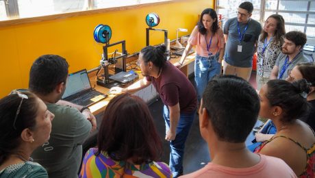 A imagem mostra um grupo de pessoas reunidas em um ambiente educacional ou de treinamento, acompanhando atentamente uma explicação sobre o uso de impressoras 3D. No centro, um homem de cabelos presos e óculos inclina-se para a frente, apontando para a tela de um notebook enquanto explica algo ao grupo. Atrás dele, há uma bancada com impressoras 3D em funcionamento, com carretéis de filamento azul montados acima delas. O ambiente tem uma parede amarela ao fundo e grandes janelas que permitem a entrada de luz natural. O grupo é composto por homens e mulheres de diferentes idades e estilos, muitos usando crachás azuis, sugerindo um evento ou capacitação. Algumas pessoas observam atentamente o instrutor, enquanto outras olham para os equipamentos. A cena transmite um clima de aprendizado, colaboração e interesse por tecnologia e inovação.