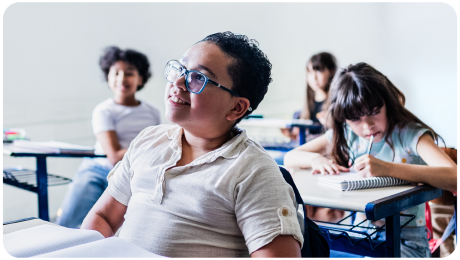 A imagem mostra uma sala de aula com crianças estudando. Em primeiro plano, um menino de óculos azuis e camisa branca está sorrindo e olhando atentamente para algo ou alguém fora do quadro. Ele está sentado em uma carteira escolar com um caderno aberto à sua frente. Ao fundo, outras crianças também estão envolvidas em atividades. À direita, uma menina de cabelos longos e franjas está concentrada, escrevendo em seu caderno com um lápis. Mais atrás, duas outras crianças, uma de cabelos cacheados e outra de cabelos lisos, estão sentadas em suas carteiras, parecendo acompanhar a aula. A sala tem paredes claras e carteiras de estrutura metálica azul, transmitindo um ambiente típico de escola. A iluminação natural destaca os rostos das crianças, dando um tom acolhedor à cena.