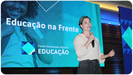 A imagem mostra uma mulher segurando um microfone e discursando em um evento. Ela veste uma blusa clara de mangas compridas e uma calça escura. Seu cabelo está preso em um coque baixo, e ela tem uma expressão confiante enquanto gesticula com a mão esquerda. Ao fundo, há um grande telão azul com a frase 