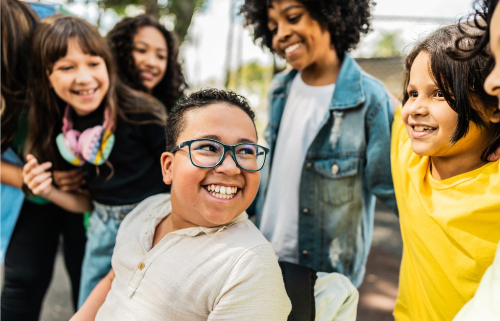 Menino cadeirante, sorridente e de óculos, está cercado por outras crianças que também sorriem e interagem com ele em ambiente externo, transmitindo inclusão e amizade.