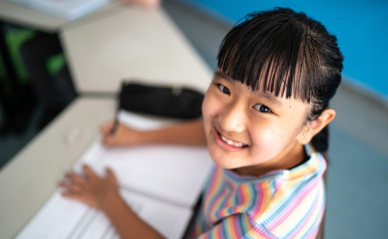 Menina sorridente, com cabelo preso e franja reta, escreve em um caderno em sala de aula. Ela usa camiseta listrada e olha para a câmera.