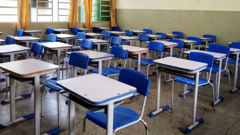 Sala de aula vazia, com várias carteiras escolares organizadas em fileiras. As carteiras têm tampo branco e cadeiras azuis, com estrutura metálica. Ao fundo, há janelas com vidros parcialmente abertos e cortinas amarelas, além de uma parede pintada em dois tons de verde e bege. O chão é de piso frio, refletindo a luz natural que entra pelas janelas.